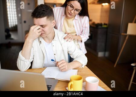 man looking frustrated, having no money to pay off debts, managing family budget together, sitting home at kitchen table with lots of papers, laptop a Stock Photo