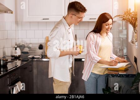 attractive cheerful girl in casual clothes preparing for exams, education, family writing a shopping list in the kitchen Stock Photo