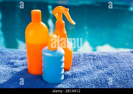 Cosmetic sunscreen products, set different orange bottles of sun protection body cream on blue towel against swimming pool. Skin care and protection Stock Photo