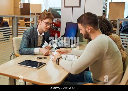 young man consulting couple at motor show, close up photo. conversation, consultation. shop assistant pointing to the document, contract. look here Stock Photo