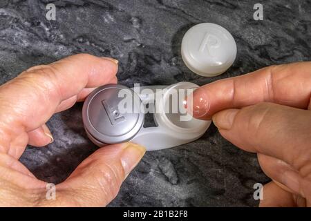 A woman’s finger with an eye contact lens on a fingertip, ready to remove from or put away in the right side of a storage pot with cleansing solution. Stock Photo