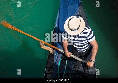 Overhead view of Venetian gondolier in traditional hat and striped shirt rowing a gondola under a bridge on a canal in Venice, Italy Stock Photo