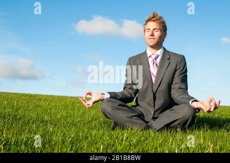 Meditating office worker sitting with eyes closed in a cross-legged yoga pose outdoors in summery green field Stock Photo