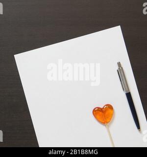 Template of white paper with pen and heart shaped caramel on dark wenge color wooden background. Concept of entertaining children's business, leisure for kids. Stock photo with empty space for text. Stock Photo