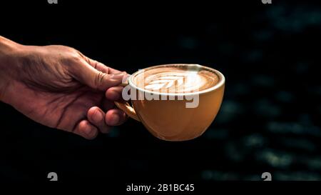 Hand Hold Cup Hot Latte Art in yellow mug against dark background. Stock Photo