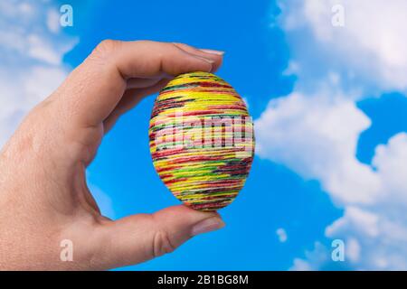 Hand holding handmade colorful yellow Easter egg on sunny spring blue sky background. Empty chicken eggshell wrapped by glued colorfully painted twine. Stock Photo