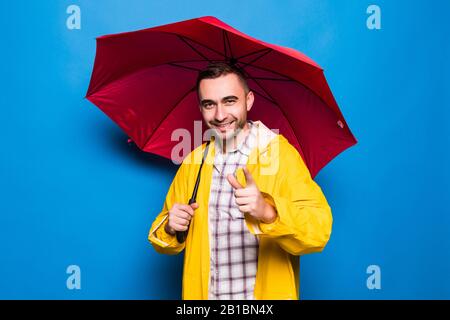 Young handsome bearded man in yellow raincoat with red umbrella pointed on you over blue background Stock Photo