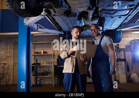 Portrait of two modern car mechanics standing under vehicle on car lift and using laptop, copy space Stock Photo