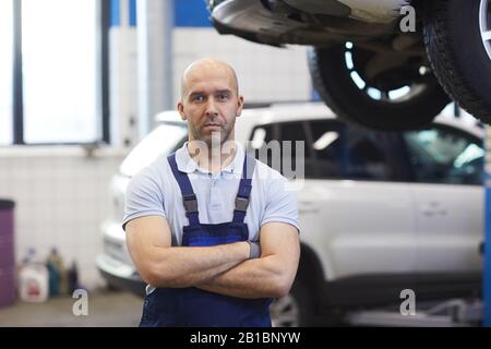 Waist up portrait of muscular car mechanic standing with arms crossed while posing in garage shop, copy space