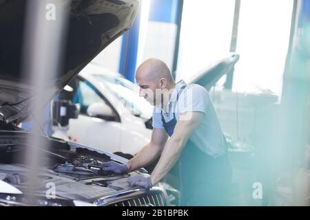 Side view portrait of muscular car mechanic looking into open hood of vehicle during inspection in garage shop, copy space Stock Photo