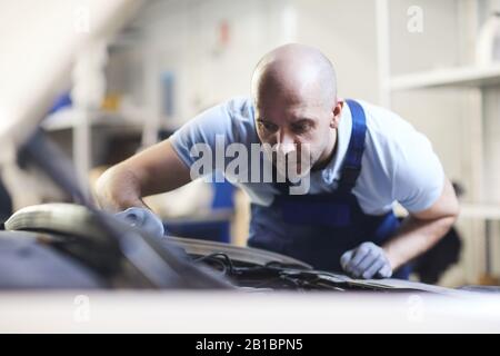 Front view portrait of muscular car mechanic looking into open hood of vehicle during inspection in garage shop, copy space Stock Photo