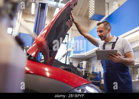 Low angle portrait of bearded car mechanic using digital tablet and opening hood during vehicle inspection in garage shop, copy space Stock Photo