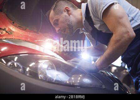 Close up portrait of bearded car mechanic looking under hood and using flashlight lamp during vehicle inspection in garage shop, copy space Stock Photo
