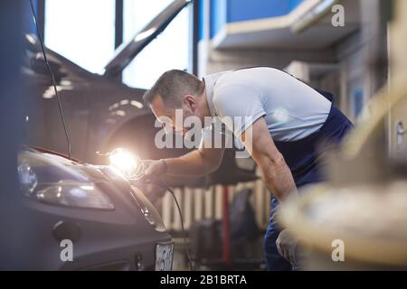 Side view portrait of bearded car mechanic looking under hood and using flashlight lamp during vehicle inspection in garage shop, copy space Stock Photo
