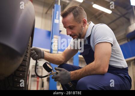 Side view portrait of car mechanic checking pressure in tires during vehicle inspection in garage shop, copy space Stock Photo