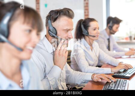 Call center worker accompanied by his team. Smiling customer support operator at work. Young employee working with a headset. Stock Photo