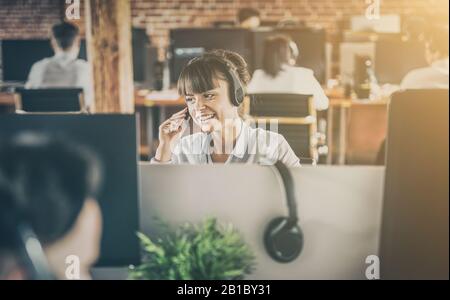 Call center worker accompanied by her team. Smiling customer support operator at work. Young employee working with a headset. Stock Photo