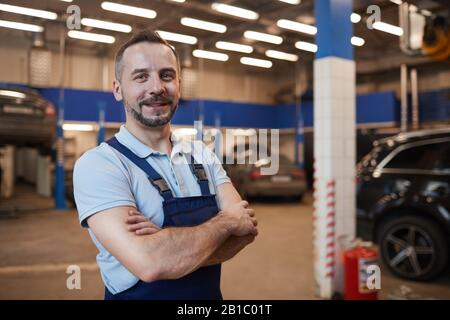 Waist up portrait of smiling car mechanic standing with arms crossed while posing in auto repair shop, copy space Stock Photo