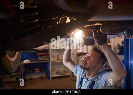 High section view at mature mechanic looking under car on lift and holding lamp light during inspection in auto repair workshop, copy space Stock Photo
