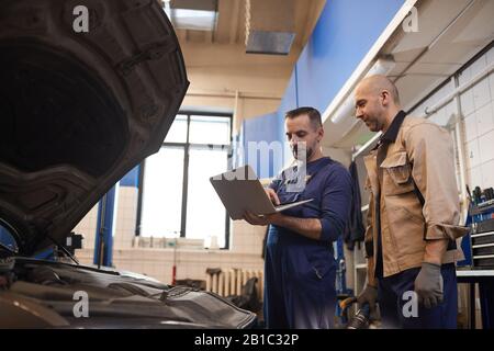 Side view portrait of two car mechanics using laptop while inspecting vehicle in auto repair workshop, copy space Stock Photo
