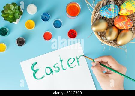 Easter concept photo, preparing for Easter basket with hand made Easter eggs next to multicolor paints and hand with brush which paint Easter on a blu Stock Photo