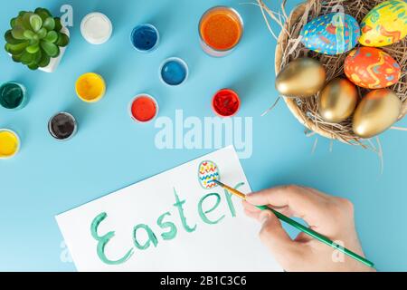 Easter concept photo, preparing for Easter basket with hand made Easter eggs next to multicolor paints and hand with brush which paint Easter egg on a Stock Photo