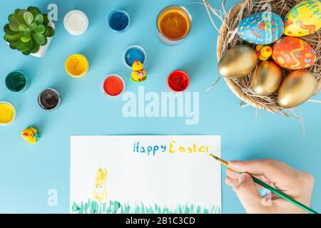 Easter concept photo, preparing for Easter basket with hand made Easter eggs next to multicolor paints and hand with brush which paint happy Easter on Stock Photo