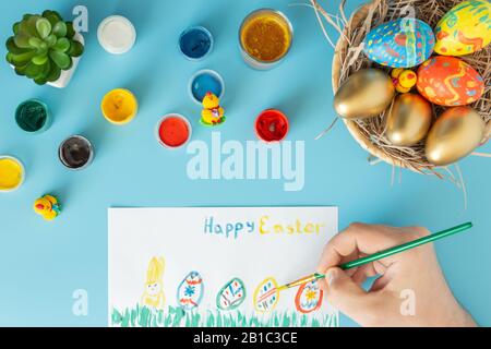 Easter concept photo, preparing for Easter basket with hand made Easter eggs next to multicolor paints and hand with brush which paint multicolor East Stock Photo