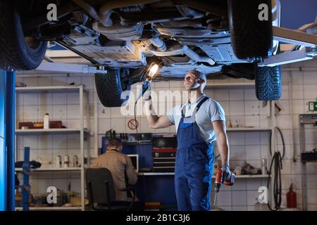 Portrait of muscular car mechanic inspecting vehicle standing under car on lift in auto repair workshop, copy space Stock Photo