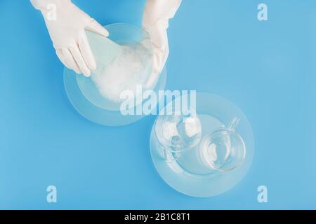 Female hands in pearly white gloves washing beautiful light blue glass plates and cups on trendy blue background. Washing dishes concept. Backdrop for Stock Photo