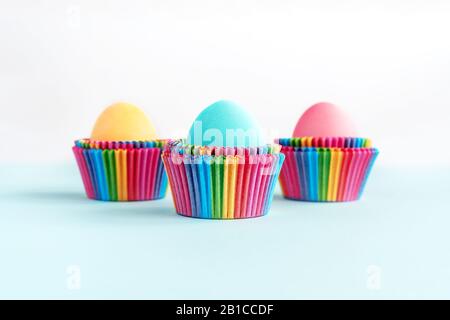 Bright creative Easter decoration of colorful eggs in raibow colored paper forms for cupcakes on white and blue background. Mockup for your design. Co Stock Photo