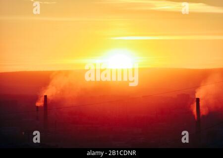 Dark harmful smoke from chimneys of power plant silhouette on sunset clear orange sky. Stock Photo