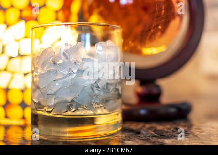 An alcohol glass of crushed ice sits aside a round container of scotch whiskey on a granite countertop with warm light Stock Photo