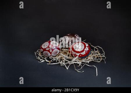 Three traditionally Lithuanian painted red, black, white, blue eggs with wax, called marguciai, in a straw on a black background. With space for text Stock Photo