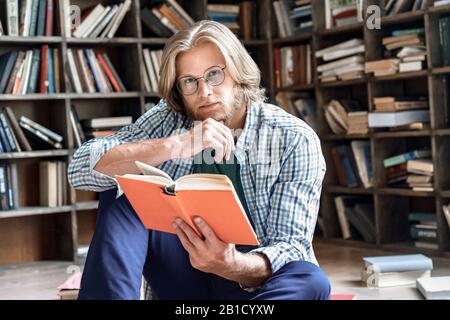 Thoughtful young male student glasses read book library floor education concept Stock Photo