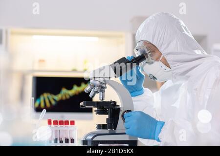 Side view of researcher in protective suit and mask using microscope while studying virus in laboratory Stock Photo