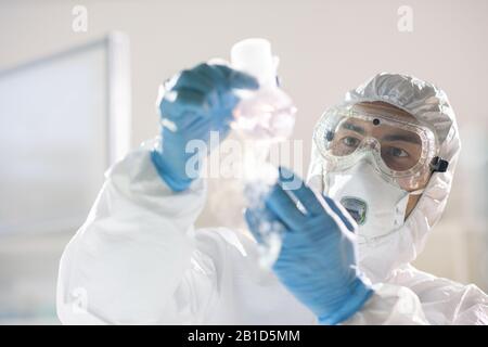 Concentrated chemical researcher in protective suit and respiratory mask holding flask with smoking substance Stock Photo