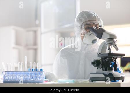 Concentrated Asian virology researcher in protective suit using microscope while testing virus sample in laboratory Stock Photo