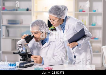 Focused scientist in safety goggles using microscope while working on medical research with Asian assistant Stock Photo