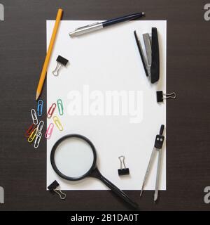 Template of white paper with pen, magnifier, stapler, paper clips and other stationery on dark wenge color wooden background. Stock photo with empty space for text  Stock Photo