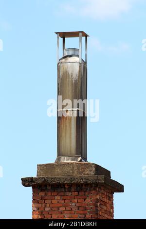 Dirty old industrial chimney with metal cap on top made of partially rusted thin metal on top of red brick foundation on clear blue sky background Stock Photo