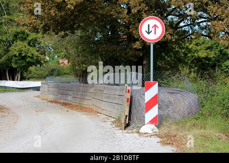 Give way to oncoming vehicles round road sign mounted on top of metal pipe with striped red and white warning sign in front of thick temporary flood Stock Photo