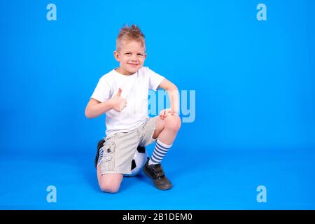 Full length Cheerful caucasian child boy shows thumbs up sign, sitting on soccer ball over blue studio background. Football game and Soccer fans Stock Photo