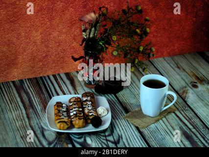 A plate with three eclairs, a cup of tea and a vase with a herbarium are on the table against the wall. Close-up. Stock Photo