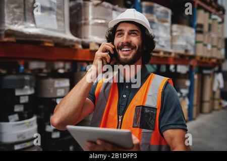 Warehouse worker using digital tablet while in conversation over mobile phone in warehouse wearing white helmet and safety vest Stock Photo