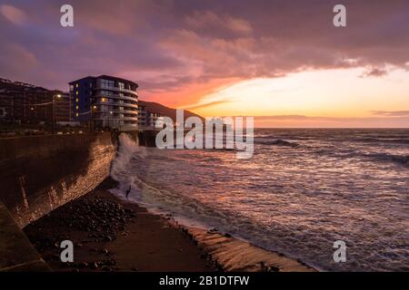 A late winter sunset over Westward Ho! Promenade in North Devon. Rough Atlantic waves crash over the seawall as it nears high tide Stock Photo