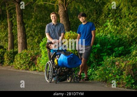 Handsome Caucasian father in early fifties walking in park outdoors with two biracial Asian Caucasian sons, one with special needs in wheelchair Stock Photo
