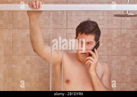 Young Caucasian man speaks on the phone in the shower. Serious white male uses a smartphone in the bathroom. Stock Photo