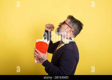 Portrait of a funny white beard man, stunned, crazy and speechless, shocked by the plot twist of the movie he is looking at holding a box of popcorn f Stock Photo