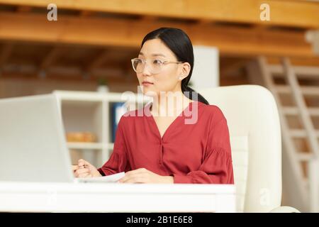 Portrait of successful Asian woman wearing red blouse working at desk in modern white office, copy space Stock Photo